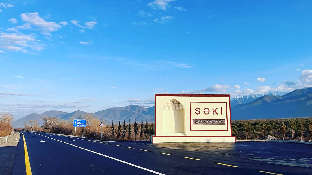 Şəki girişindəki lövhə və arxada qarlı Böyük Qafqaz dağları